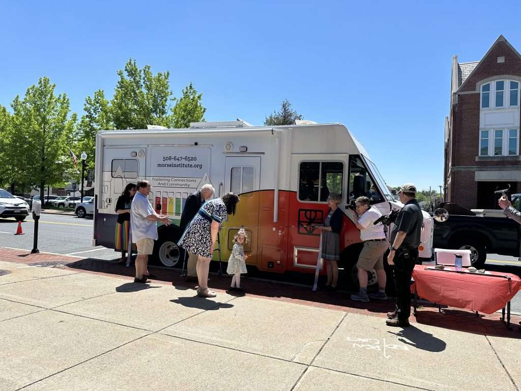 In front of a bookmobile decorated with a picture of the Morse Institute Library and rainbow colors, Dr. Keefe and his great-granddaughter prepare to cut a ribbon while others look on excitedly. 