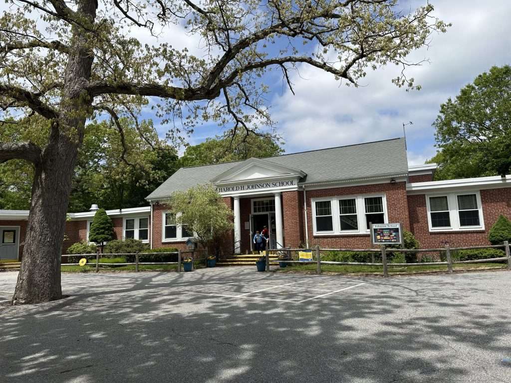 A brick elementary school with white trim. There is a tree and parking lot in front of the school. The school’s door is open, and two students are running out.