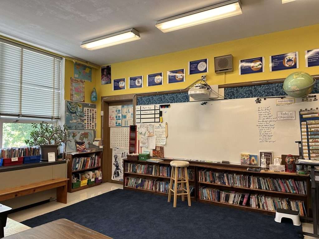 A homey classroom with yellow walls, a blue rug, and wooden shelves full of books.