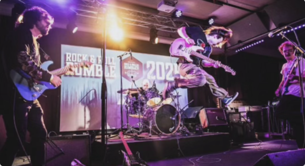 Danahy is a couple feet in the air while strumming his guitar. He is onstage with two bandmates. There is purple lighting and a backdrop that reads "Rock&Roll Rumble 2024."