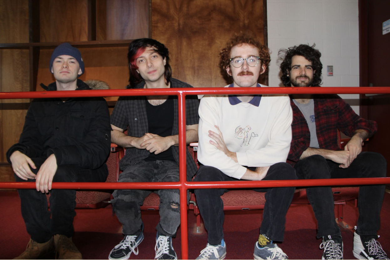The four band members sit on red seats in front of a wooden wall.