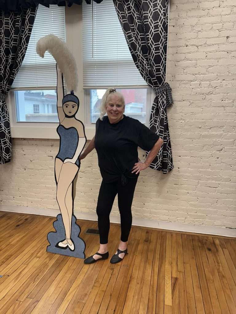 Nancy Kelley stands in her studio next to a life-size wooden decoration of a dancer with a large feather on its hat.