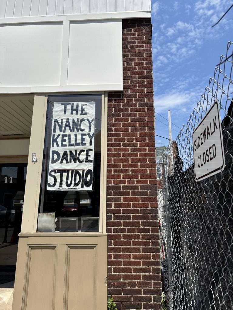 A handmade "Then Nancy Kelley Dance Studio" sign on a brick wall next to a "Sidewalk Closed" sign on the fence surrounding the construction zone.