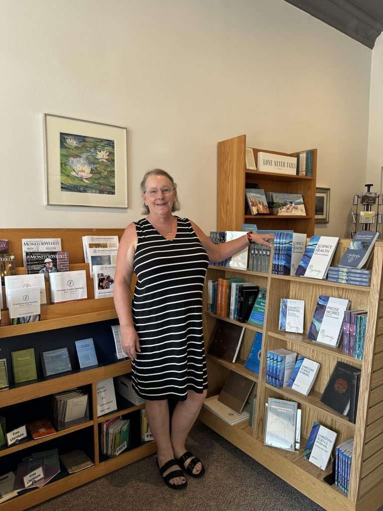 Linda Zanoni stands between two bookshelves holding Christian Science books and publications. A "Love Never Fails" sign is visible in the background on another bookshelf.