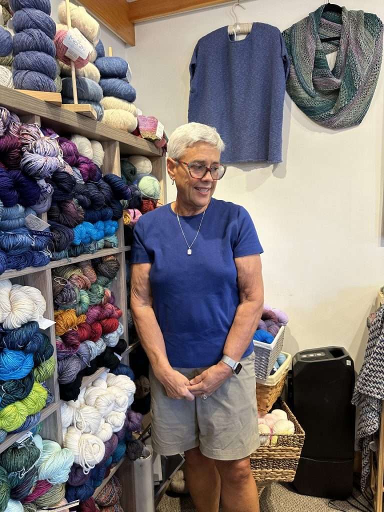 With a slightly worried look on her face, Deborah Smith stands next to a large shelf filled with many colors of yarn in Iron Horse.