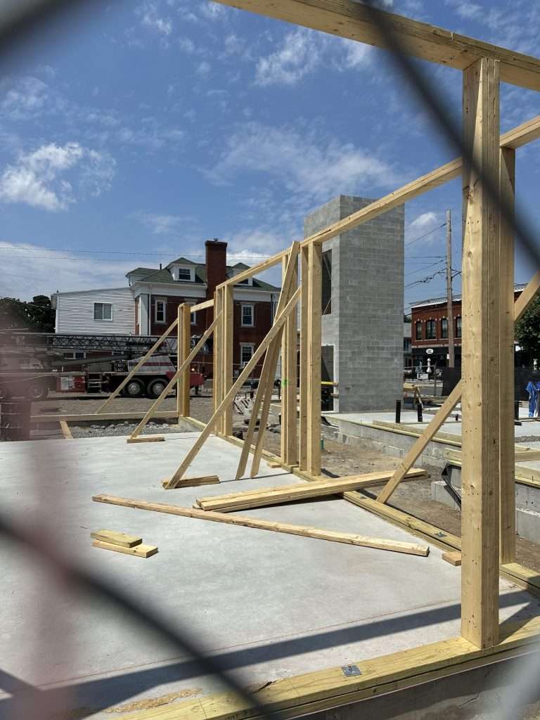 Cement foundations, wooden beams, and what appears to be an elevator shaft are visible through the fence surrounding the construction site.